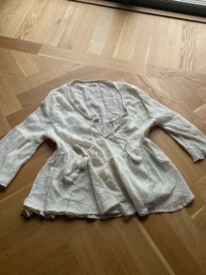 Vit boho-blus med knytband - Säljer en luftig vit blus i boho-stil med trekvartsärm och dekorativa knytband med tofsar framtill. Blusen har lätt struktur i tyget och en avslappnad passform.