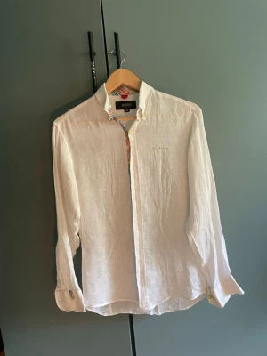 Vit linneskjorta från Dahlin XS - Säljer en vit långärmad skjorta från Dahlin i svalt linne. Skjortan har klassisk krage, knappar framtill och diskret broderad logga på bröstet. Perfekt för en clean och fräsch stil. Passar dig som gillar minimalistiskt och stilrent.