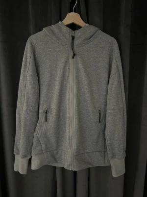 Grå CP company tröja  - Säljer en grå hoodie med hel dragkedja, två fickor med dragkedja och en unik huva med inbyggda glasögon. Hoodien har ribbade muddar vid ärmar och nederkant. Perfekt för dig som vill sticka ut med en cool detalj på huvan.