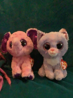Två TY Beanie Boo gosedjur - Säljer två mjuka TY Beanie Boo gosedjur, en rosa elefant med lila öron och en ljuslila katt med vita detaljer och rosa nos. Båda har stora glittriga ögon och är perfekta som samlarprylar eller dekoration. Taggarna sitter kvar.