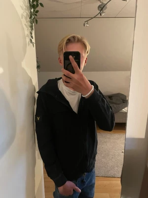 Lyle & Scott hoodie  - Säljer en svart Lyle and Scott hoodie med hel-zip och huva. Den har en bröstficka och en gul broderad logga på armen. Långa ärmar och ribbade muddar vid ärmslut och nederkant. Inga defekter. Perfekt för chill dagar eller när du vill ha en enkel och clean look. Storlek = 12/13 år vilket motsvarar 158-164cm. Hör av er vid frågor! 