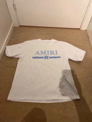 Vit t-shirt från Amiri med blått tryck - Säljer en vit t-shirt från Amiri med ljusblått tryck på bröstet. T-shirten har klassisk passform, rund halsringning och korta ärmar. Perfekt för dig som gillar stilrena och trendiga plagg.