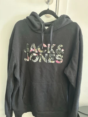 Svart hoodie från Jack & Jones - Svart hoodie från Jack & Jones med stor logga i blommigt mönster. Lite nopprig men går att få bort.