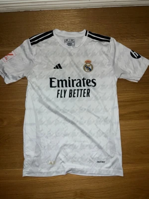 Real Madrid matchtröja Mbappé nr 9 - Säljer en vit Real Madrid matchtröja från Adidas med svarta detaljer och tryck. Framsidan har klubbmärke och sponsorlogga, baksidan har Mbappé och nummer 9. Tillverkad i lätt och ventilerande material med HEAT.RDY-teknologi.