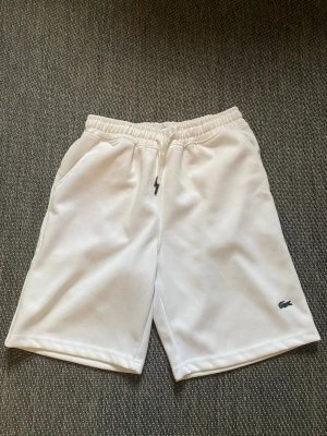 Vita shorts från Lacoste med  - Säljer ett par klassiska vita shorts från Lacoste med elastisk midja och sidofickor. Shortsen har en clean look med den ikoniska krokodilloggan broderad på benet. Perfekta för sommaren och sportiga outfits.