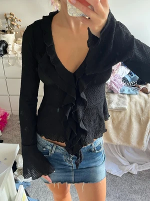 Svart blus med volang och v-ringning - Säljer en svart blus med långärmad design och djup v-ringning. Blusen har volangdetaljer längs halsringningen och framtill, samt lätt utsvängda ärmslut. Materialet är strukturerat och ger en trendig look. Helt oanvänd så mycket gott skick.