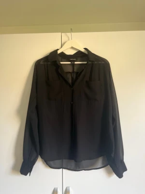 Svart transparent blus från Monki - Snygg svart blus från Monki i transparent material. Blusen har långa ärmar, klassisk krage och knäppning framtill. Två bröstfickor och avslappnad passform ger en cool och trendig look. Perfekt att styla lager på lager.