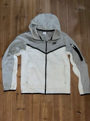 Grå och vit Nike Teach Fleece - Nike hoodie i grått och vitt med hel dragkedja framtill, svart Nike-logga på bröstet och svarta detaljer. Huva och fickor på sidorna samt en extra ficka med dragkedja på ena ärmen. Tillverkad i mjukt och stretchigt material. En sällsynt Nike tech fleece limited edition som inte säljs längre! Knappt använd då de inte var min stil!  Nypris:1559kr.   Mitt pris:845kr
