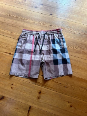 Burberry shorts - Snygga shorts med klassiskt rutigt mönster i beige, svart, vitt och rött. Shortsen har elastisk midja med snörning och sidofickor samt en bakficka. Perfekta för sommaren och ger en trendig look.