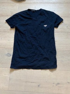 Mörkblå t-shirt med v-ringning - Snygg mörkblå t-shirt med v-ringning och ett stilrent emblem på bröstet. T-shirten har korta ärmar och är tillverkad i mjuk bomull, perfekt för en avslappnad look.