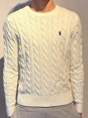 Vit oanvänd Polo Ralph Lauren stickad - Kabelstickad vit tröja från Polo Ralph Lauren med rund halsringning och diskret broderad logga på bröstet. Tröjan har ribbade muddar vid ärmslut och nederkant, och är tillverkad i mjukt material som ger en klassisk och clean look.