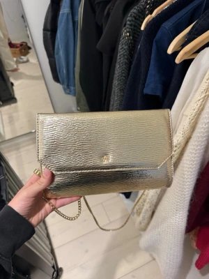 Guldig kuvertväska med kedja - Snygg kuvertväska i metallic guld med struktur och stilren design. Väskan har en tunn guldfärgad kedja som axelrem och magnetlås. Perfekt för att lyfta din outfit med lite extra bling. Rymmer mobil, kort och småsaker.