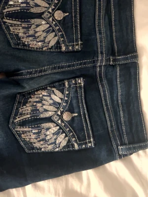 Mörkblå jeans med fickor - Snygga mörkblå jeans med detaljerade bakfickor som har silverfärgade broderier och paljetter i fjädermönster. Jeansen har kontrastsömmar och dekorativa knappar på fickorna. Perfekta för dig som vill sticka ut lite extra.