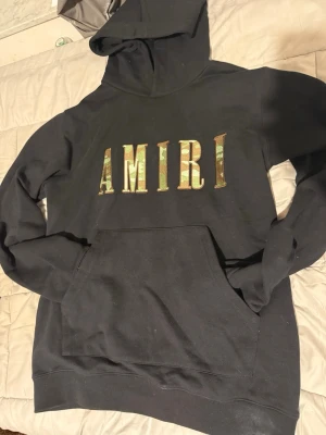 Svart hoodie från Amiri med camotryck - Säljer en svart hoodie från Amiri i storlek Large. Framsidan har stort AMIRI-tryck i camomönster. Klassisk känguruficka och huva. Perfekt för dig som gillar streetwear och vill sticka ut med en snygg detalj på bröstet.