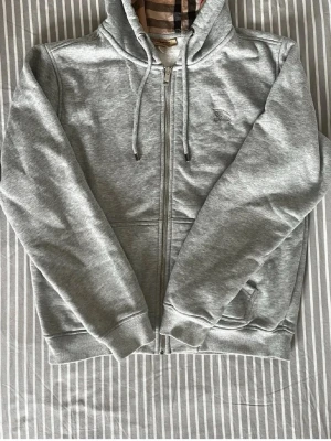 Grå burberry  zip hoodie - Snygg grå hoodie med dragkedja och huva med dragsnören. Broderad logga på bröstet och ribbade muddar vid ärmslut och nederkant. Tillverkad i mjukt sweatshirtmaterial, perfekt för en avslappnad stil.