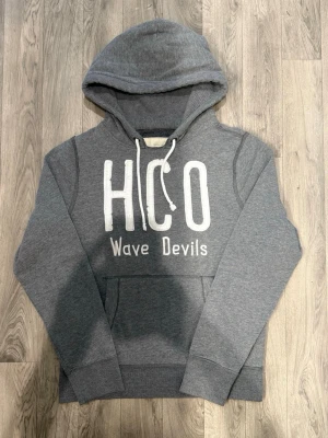 Hollister Wave Devils Hoodie Grå Strl XL (Passar L) - Hollister Wave Devils grå hoodie med vit HCO-logga framtill, märkt storlek XL men passar bättre som en normal L. Axelbredd 44 cm, bröstvidd 58 cm, armlängd 70 cm, midja 48,5 cm, längd 68 cm, i fint skick.