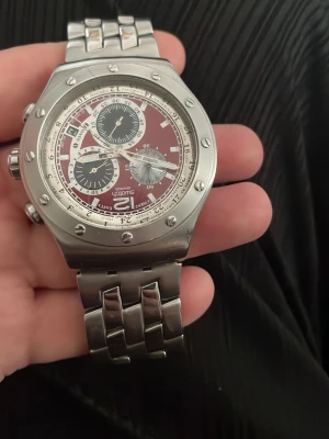 Swatch kronograf i rostfritt stål - Snygg Swatch kronograf med vinröd urtavla och flera subdials. Boetten är i borstat rostfritt stål med markerade skruvar och armbandet är i matchande metall. Klockan har datumvisning och tydliga indexmarkeringar. Perfekt för dig som gillar sportig och modern stil.