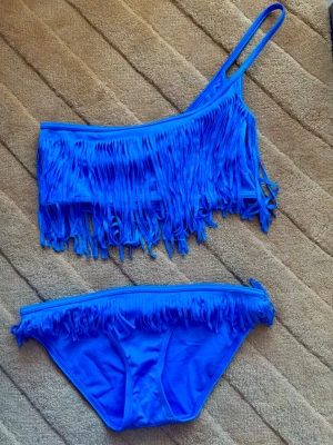 Ny Asos Klarblå bikini med fransar och asymmetrisk topp - Säljer en oanvänd djupblå bikini med en axelband och fransar både på toppen och trosan. Toppen har en asymmetrisk design som ger en unik look. Perfekt för sommaren och strandhäng! Materialet känns mjukt och stretchigt. Storlek 36 (trosa) och 38 (topp). Se gärna mina övriga annonser för bundlerabatter och samfrakt!