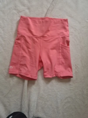 Rosa träningstights shorts - Snygga rosa träningstights shorts med hög midja och praktiska sidofickor. Tillverkade i stretchigt syntetmaterial som passar perfekt för aktiva dagar. Märket 'aim'n' syns diskret på benet. Köpta på vinted men aldrig använda av mig bara prövade 💕 be om bilder på! Dom är lite mer corall i verkligheten!