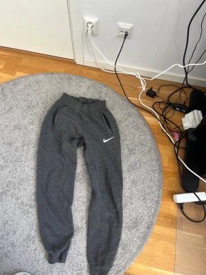 Mörkgråa Nike mjukisbyxor - Säljer ett par mörkgråa mjukisbyxor från Nike med resår i midjan och muddar vid bensluten. Byxorna har två fickor med dragkedja och den klassiska Nike-loggan broderad på ena benet. Perfekta för chill eller träning.