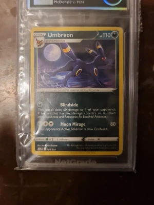 Pokémonkort Umbreon holografisk grade 9 - Säljer ett holografiskt Pokémonkort med Umbreon-motiv. Kortet har en mörkblå och gul ram, och Umbreon syns mot en nattlig bakgrund med fullmåne. Kortet är inplastat i ett skyddande case och har tydliga samlardetaljer.