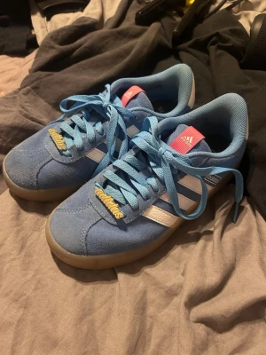 Blå Adidas sneakers med vita detaljer - Snygga blå sneakers från Adidas med klassiska vita ränder på sidorna, ljusblå skosnören och gummisula. Skorna har rosa detaljer på plösen och hälflik samt Adidas-logga i guld på snörningen. Ovandelen är i mocka och textil.