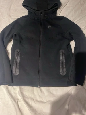 Svart Nike hoodie med dragkedja - Om du har XS kommer tröjan sitta fint ja tycker den sitter perfekt Snygg svart hoodie från Nike med hel dragkedja framtill och två fickor med dragkedja. Klassisk Nike-logga på bröstet och huva för extra stil och komfort. Perfekt till chill eller streetwear. Materialet känns mjukt och följsamt. Dragkedjan har tappat lite färg men annars är den som ny knappt använd.storlek 147-158 cm passar Xs. Köpt från JD sport.