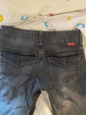 Grå bootcut jeans med ficklock - Snygga grå jeans med bootcut passform och ficklock med knapp bak. Klassisk femficksmodell och bälteshällor. Jeansen har en tvättad look och är perfekta för en avslappnad stil. Märket Angelic syns på bakfickan. Low waist