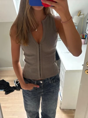 Beige ribbad ärmlös topp - Säljer en beige ribbad topp med v-ringning och dragkedja framtill. Toppen är ärmlös och har en slim passform som framhäver figuren. Perfekt att styla med jeans eller kjol för en trendig look.