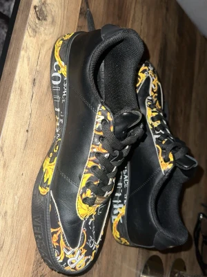 Versace Jeans Couture svarta sneakers 43 - Säljer ett par svarta sneakers från Versace Jeans Couture med färgstarka gula och orange barockmönster samt vita logotexter på sidor och sula. Skorna har svarta snören och detaljer i syntetmaterial. Perfekta för dig som vill sticka ut med din stil.