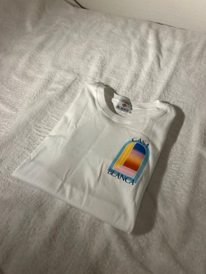Vit t-shirt från Casablanca - Snygg vit t-shirt från Casablanca med färgglad grafisk print på bröstet i blått, gult och orange. Klassisk rund halsringning och mjukt material som känns skönt mot huden. Perfekt för dig som gillar stilrena plagg med en twist.