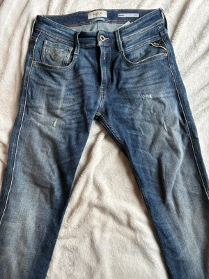 Blå slim fit jeans från Replay - Snygga blå slim fit jeans från Replay, modell Anbass. Jeansen har slitna detaljer, fem fickor och klassisk knappgylf. Tillverkade i denim med kontrastsömmar och en liten Replay-logga vid myntfickan. Perfekta för en trendig och avslappnad stil.