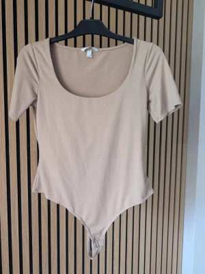 Beige basic body med kort ärm - En enkel och stilren beige body med korta ärmar och rundad urringning. Tillverkad i mjukt stretchigt material som sitter skönt mot kroppen. Perfekt att matcha med jeans eller kjol för en clean look.