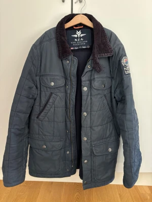 Mörkblå quiltad jacka NZ Auckland - Snygg mörkblå quiltad jacka från New Zealand Auckland med fyra framfickor och tryckknappar. Jackan har en krage i manchestertyg och patch med logga på ena ärmen. Perfekt för kyligare dagar och har en klassisk, tidlös look.