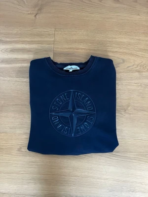 Mörkblå sweatshirt från Stone Island - Mörkblå sweatshirt med rund hals från Stone Island. Tröjan har ett stort, ton-i-ton broderat Stone Island-logotyp på bröstet och ribbade muddar vid ärmslut och nederkant. Perfekt för en avslappnad och stilren look.