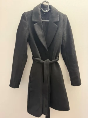 Svart kappa med knytskärp - Stilren svart kappa med klassisk krage och knytskärp i midjan. Kappan har lång ärm och rak passform, perfekt för kyligare dagar. Materialet ser ut att vara ullblandning för extra värme och komfort. Enkel och tidlös design som passar till många olika stilar.