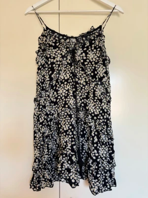Svart blommig sommarklänning från Zara, storlek XS - Söt svart klänning med vita blommor och tunna axelband. Klänningen har volangdetaljer och tyget är viskos. Modellen är kort med en ledig passform. Jättefint skick!