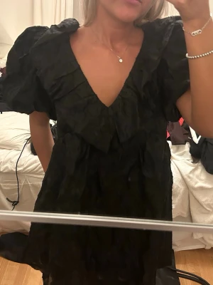 Svart kortklänning med puffärm - Säljer en svart kortklänning med djup v-ringning och stora puffärmar. Klänningen har en unik struktur i tyget och en bred krage som ger en cool look. Perfekt för dig som vill sticka ut lite extra.