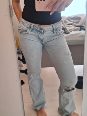Ljusblå slitna bootcut jeans - Säljer ett par ljusblå bootcut jeans med slitningar och hål vid knäna. Jeansen har klassisk femficksdesign, låg midja och är tillverkade i denim med en avslappnad passform. Perfekta för en trendig och avslappnad stil.