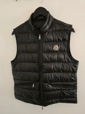 Svart Moncler gui storlek 2  - Snygg 100% äkta svart dunväst från Moncler med hög krage och två dragkedjeförsedda fickor framtill. Västen har Moncler-logga på bröstet och quiltad design. Tillverkad i glansigt material med dunfyllning för extra värme. Perfekt för lager-på-lager-stil.