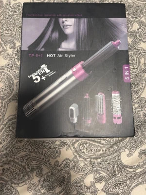 5-i-1 Hot Air Styler hårverktyg - Multifunktionellt hårstylingverktyg med fem utbytbara tillbehör för att torka, locka och borsta håret. Verktyget är silverfärgat med lila detaljer och har både rundborste och olika locktillbehör. Perfekt för att skapa olika frisyrer hemma.