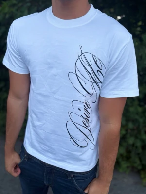 Steeze T-shirt - Snygg vit t-shirt med ett stort svart textprint i snirklig stil längs framsidan. T-shirten har rund halsringning och korta ärmar. Tillverkad i mjukt material som känns skönt mot huden. Perfekt för en clean och modern look.