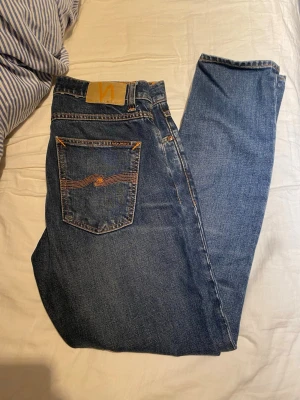 Blå jeans från Nudie Jeans - Säljer ett par klassiska blå jeans från Nudie Jeans med orange sömmar och femficksmodell. 