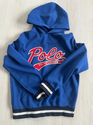 Blå hoodie från Polo Ralph Lauren - Snygg blå hoodie från Polo Ralph Lauren med stort rött och silver logotryck på bröstet. Tröjan har huva och ribbade muddar med svarta och vita ränder vid ärmslut och nederkant. Perfekt för en avslappnad och sportig stil.