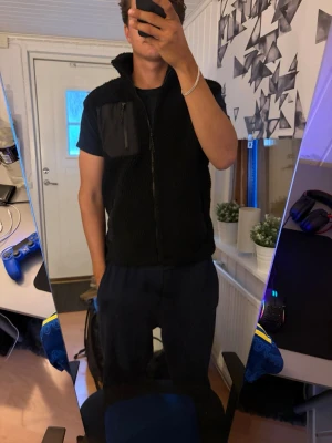 Svart fleeceväst med dragkedja - Säljer en svart fleeceväst med hög krage och dragkedja framtill. Västen har en bröstficka i nylonmaterial med dragkedja samt två sidofickor med nylonkanter. Perfekt för lager-på-lager och riktigt skön att ha på sig.