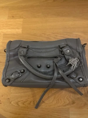 Grå handväska med detaljer - Snygg grå handväska i läderlook med silverfärgade metalldetaljer och dragkedja framtill. Väskan har både handtag och avtagbar axelrem, samt dekorativa spännen och långa läderband. Perfekt för dig som gillar en cool och trendig stil.