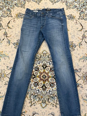 Blåa slim fit jeans med klassisk tvätt - Snygga blå jeans från Jack and Jones med slim fit passform och klassisk tvätt. Jeansen har fem fickor, bälteshällor och subtila slitningar framtill för en avslappnad look. Materialet är denim i bomull och de har dekorativa sömmar på bakfickorna. Storlek 30/30