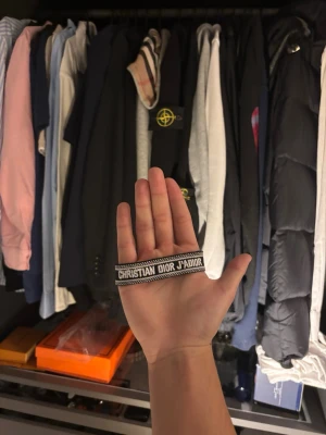 Christian Dior armband - Snyggt armband från Christian Dior med texten 'J'Adior' i vitt på svart bakgrund. Armbandet är vävt i tyg och har en stilren, modern design som passar perfekt till din outfit.
