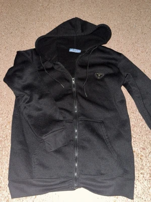 Svart hoodie med dragkedja och luva - Säljer en svart hoodie med dragkedja framtill och luva. Hoodien har fickor på sidorna och ett diskret trekantigt märke på bröstet från Prada. Muddar vid ärmslut och nederkant. Perfekt för en chill och avslappnad stil.