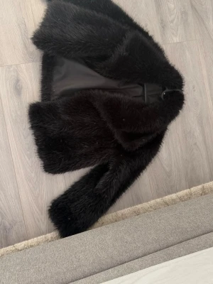 Svart fluffig pälsjacka - Säljer en svart, kort pälsjacka med lång ärm och mjukt foder. Jackan har en klassisk krage och stängs framtill med en enkel knäppning. Perfekt för dig som vill ha en trendig och varm look under kalla dagar.